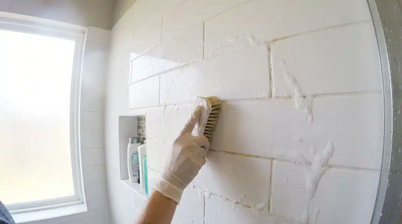 Close up of hands with a scrubbing brush cleaning grout lines on white bathroom shower tiles, a key step when you deep clean a bathroom thoroughly