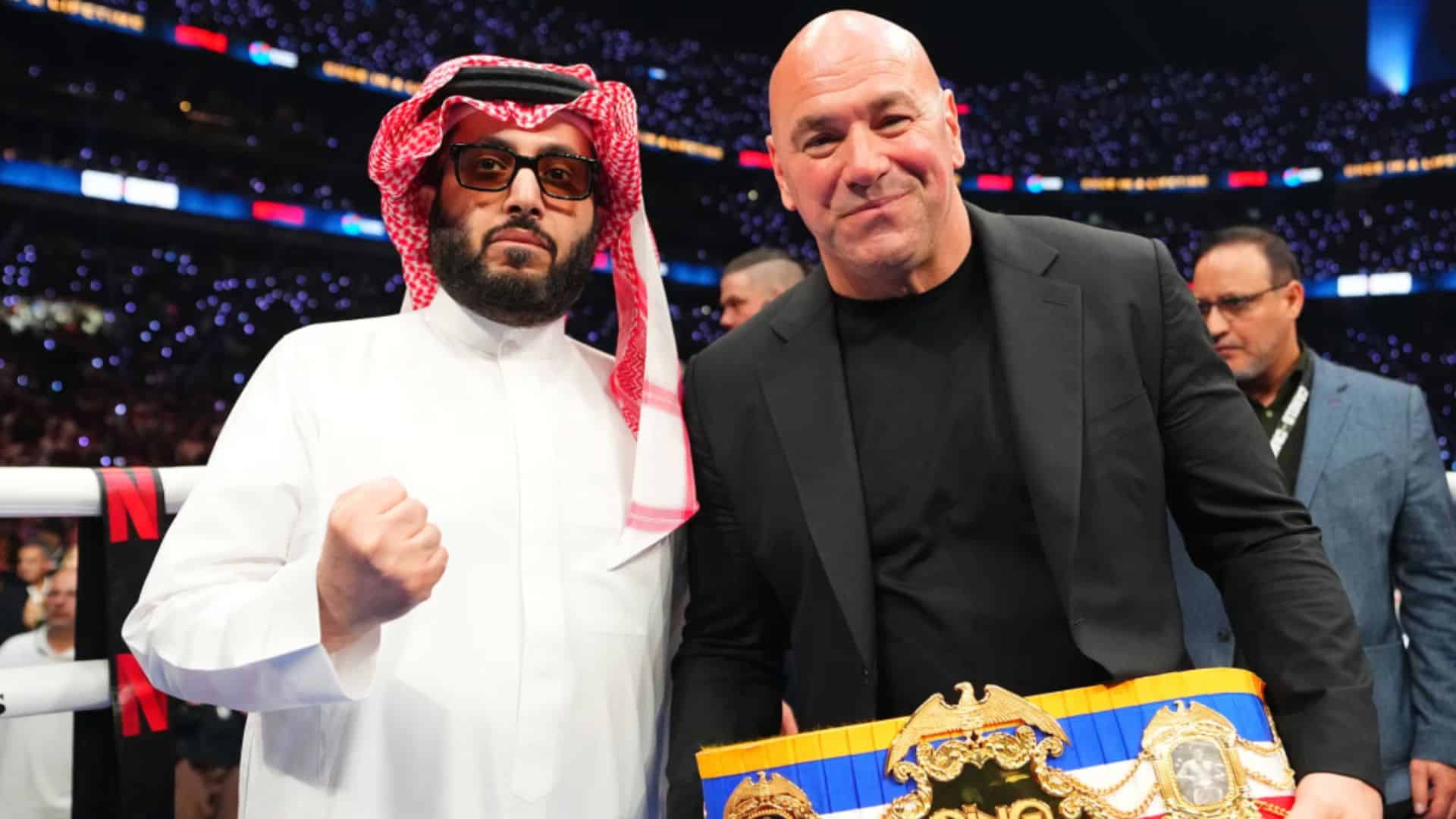 Combat sports executive posing beside a man in traditional Saudi attire while holding a championship belt.