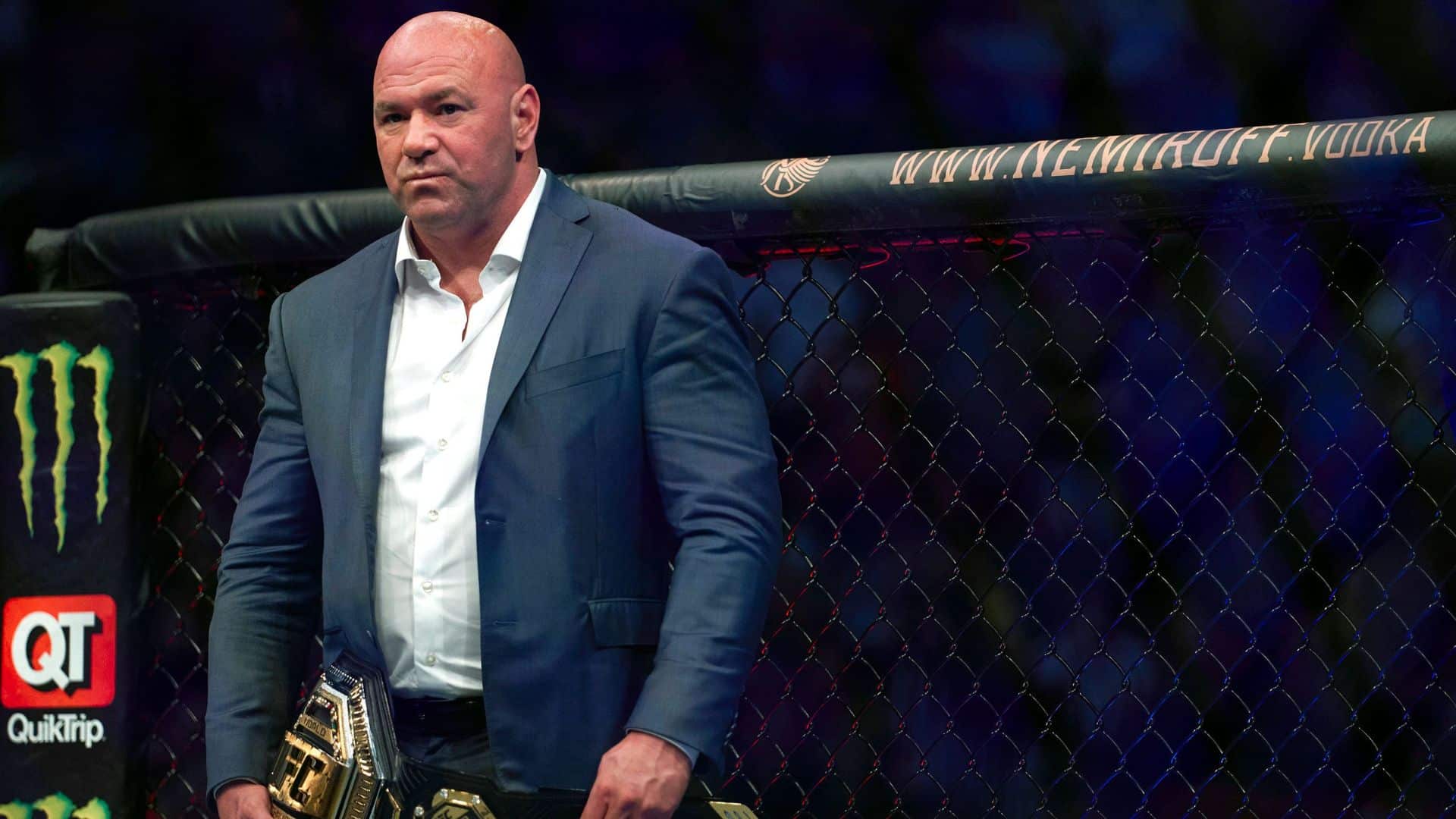 Dana white in a dark suit, standing inside an MMA octagon while holding a championship belt.