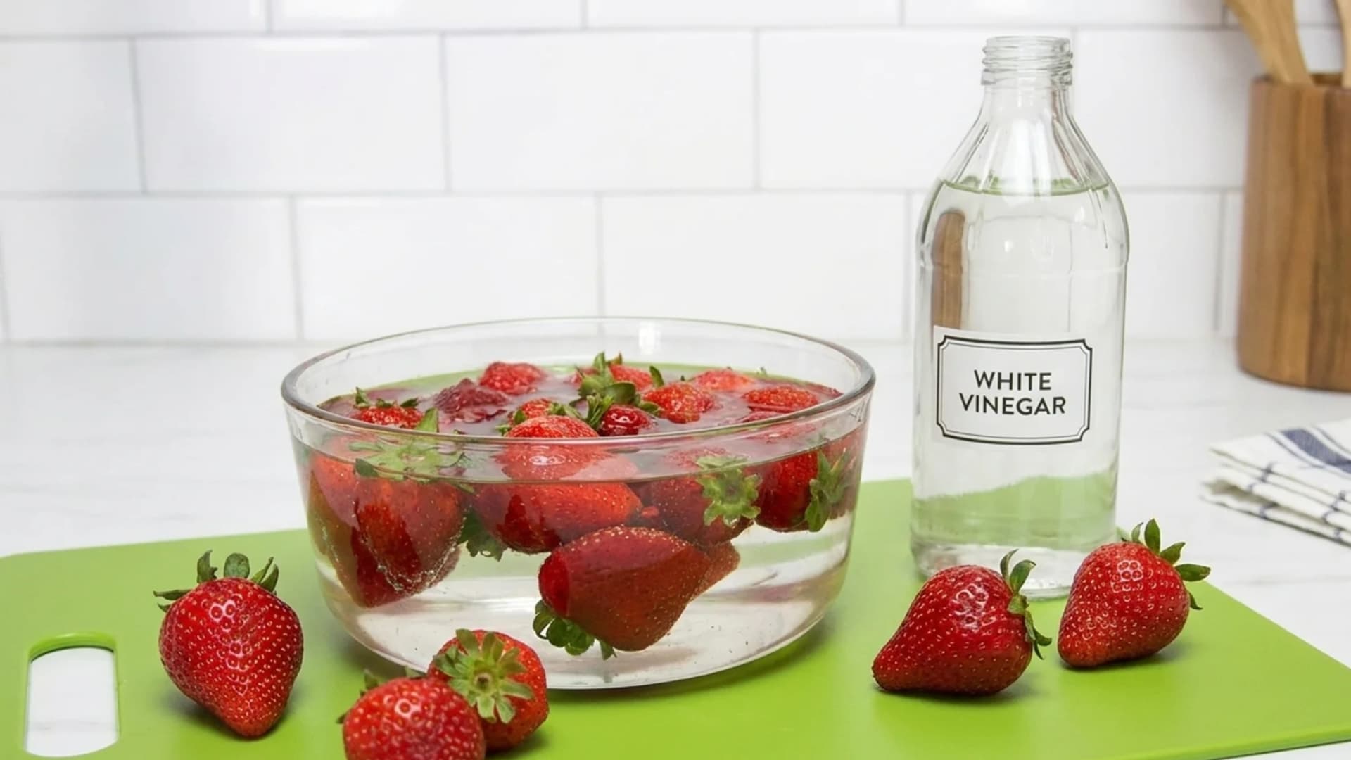 Fresh whole strawberries in a glass bowl filled with a vinegar-water bath next to a glass bottle of white vinegar on a green cutting board.