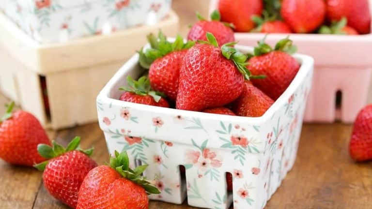 Fresh, ripe strawberries in a floral white bowl on a wooden table with additional berries in the background.