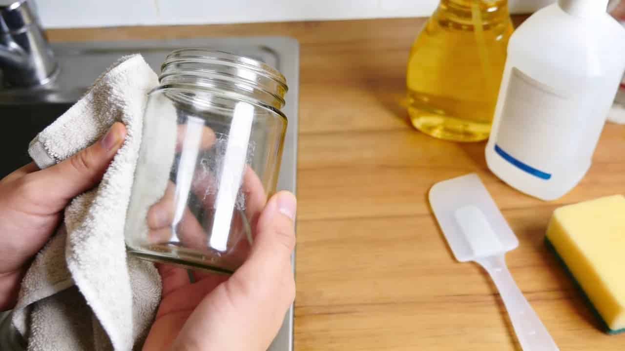 Hands removing adhesive residue from a glass jar with cleaning tools for how to remove adhesives from surfaces