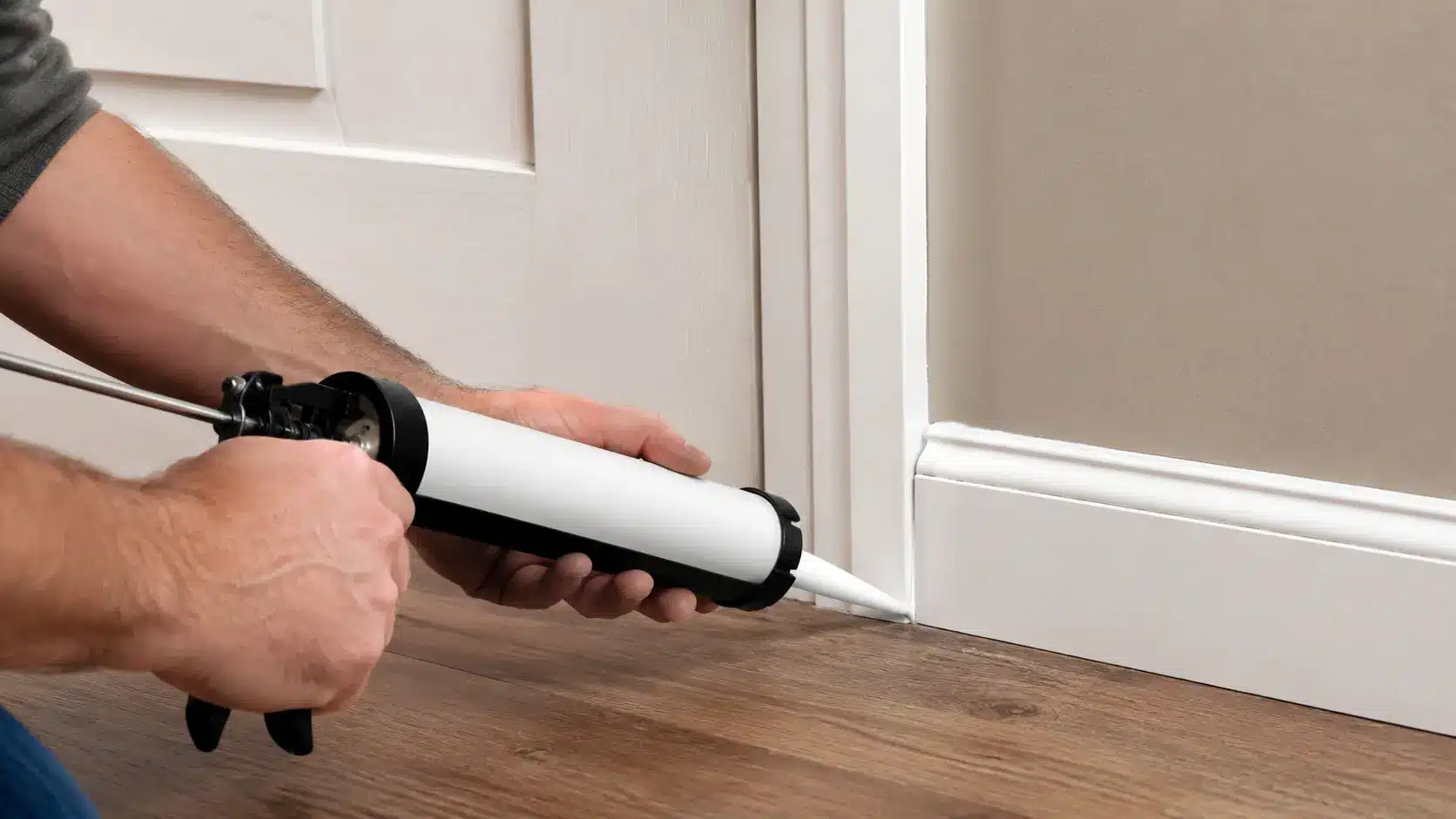 Person applying acoustic caulk along a door frame and baseboard to seal gaps and reduce sound.