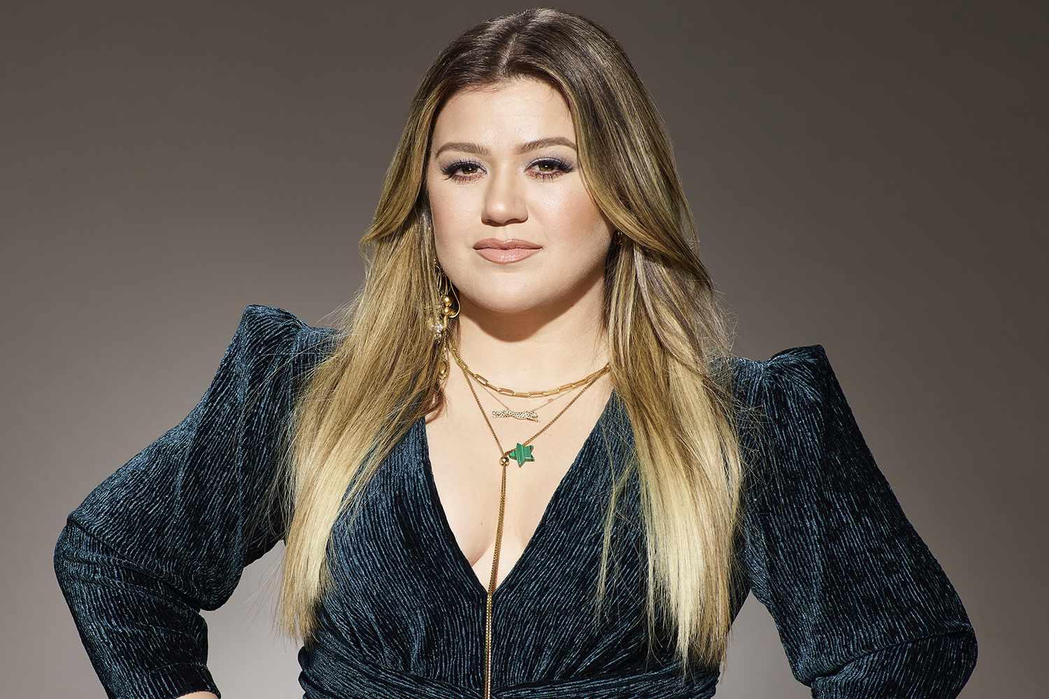 Kelly Clarkson wearing a long-sleeved teal velvet top and gold necklaces, posing for a portrait against a neutral backdrop.