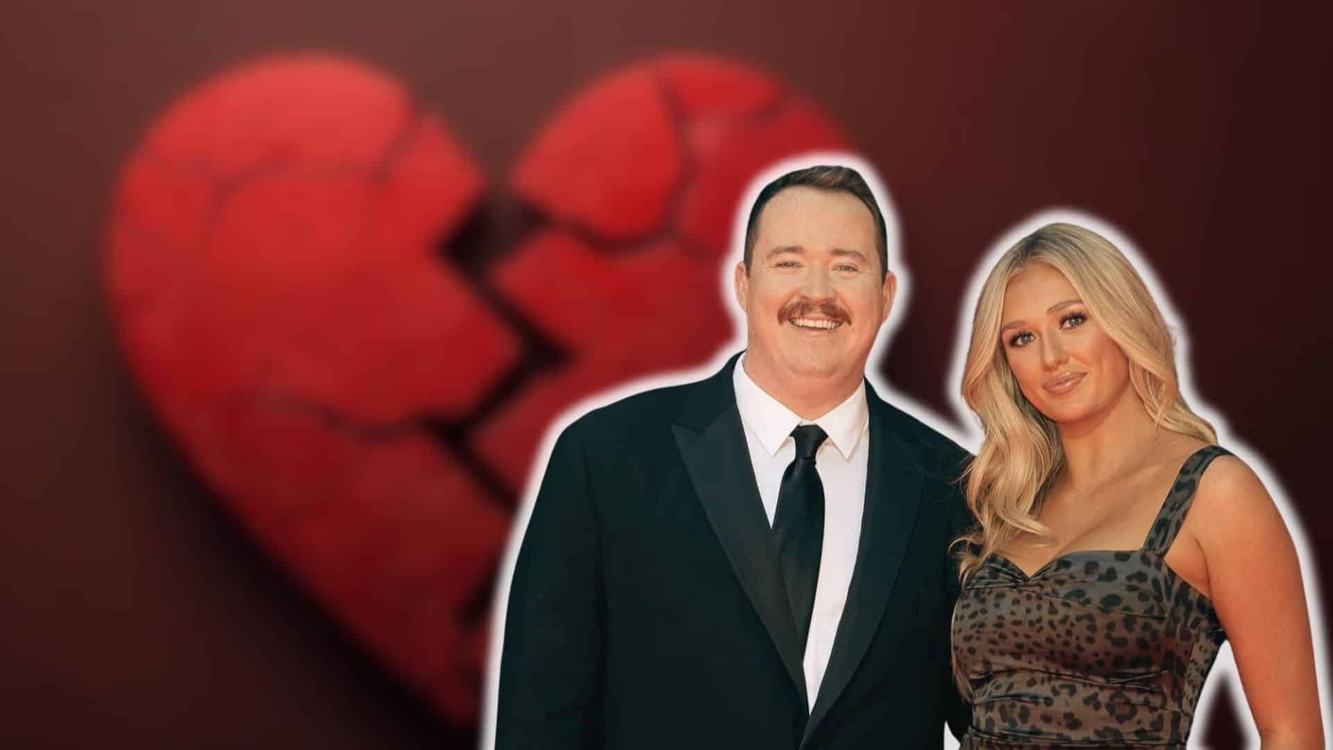 Shane Gillis posing with a blonde woman in front of a broken heart background.