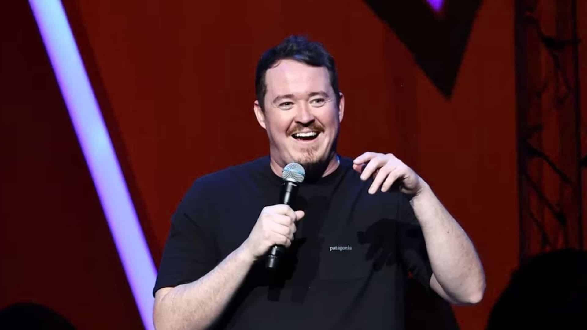 Shane Gillis smiling and holding a microphone during a stand-up comedy performance