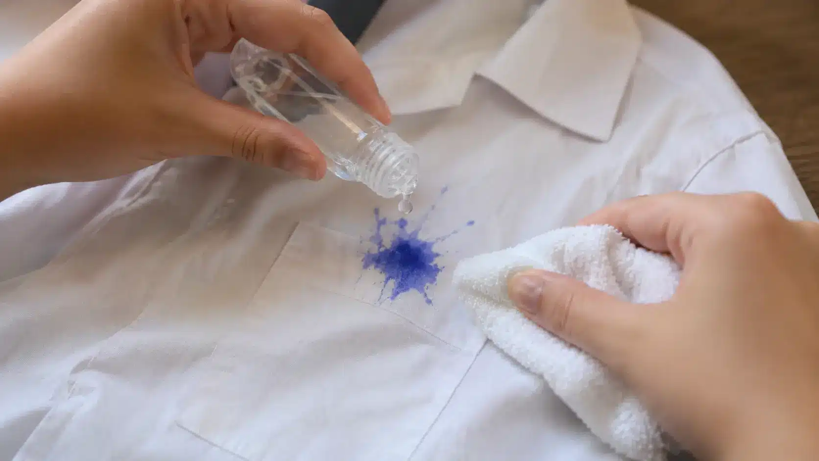 Person applying clear hand sanitizer to a blue ink stain on a white shirt while holding a clean cloth.