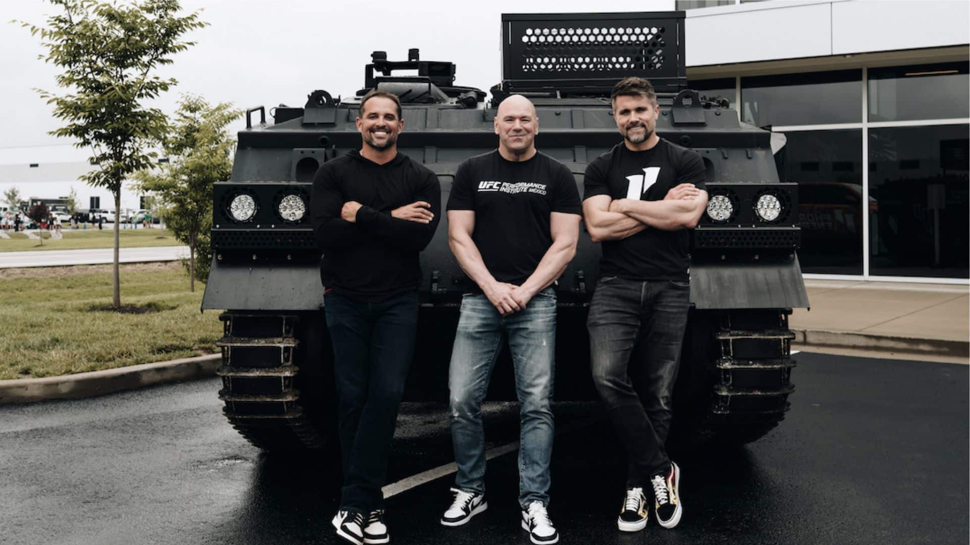 Three men posing together in front of a large armored vehicle outside a sports facility.