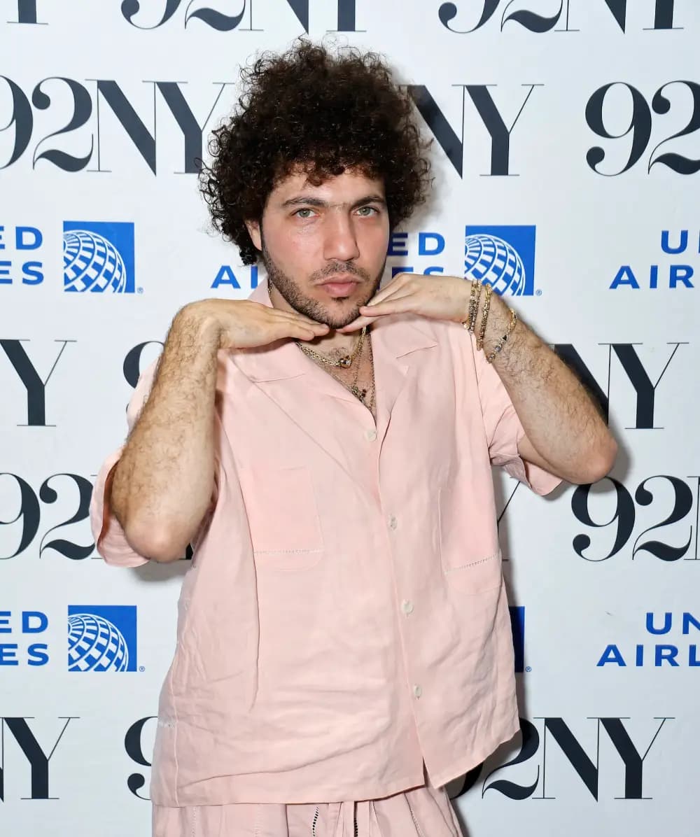 Benny Blanco posing on a red carpet in a pink button-down shirt and matching shorts