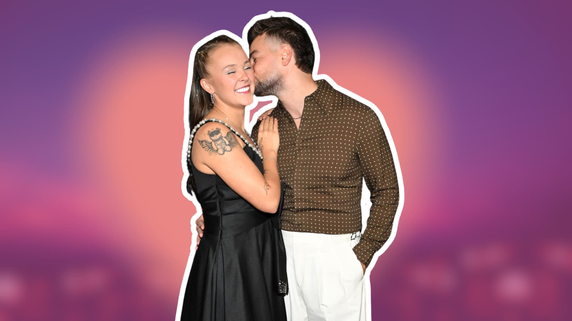 Jojo Siwa in a black dress smiles as her partner Chris Hughes kisses her cheek against a purple and pink background.
