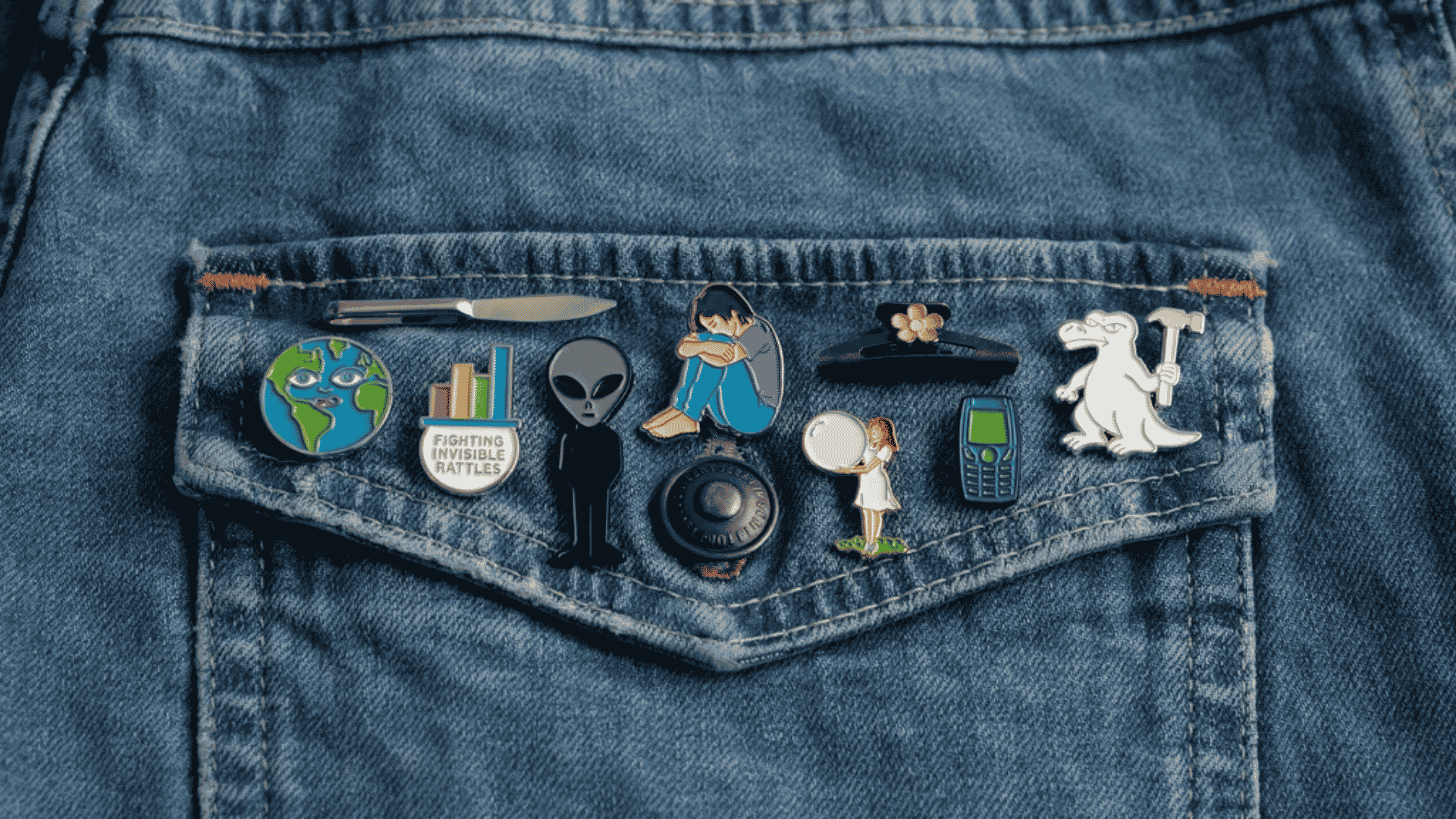 A denim jacket pocket decorated with a collection of colorful enamel pins, including space, dinosaur, and figure designs.