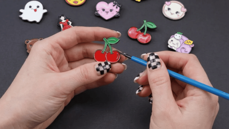 A person applying shiny clear glaze with a paintbrush to a cherry-shaped pin, surrounded by handmade enamel pins on a dark surface.