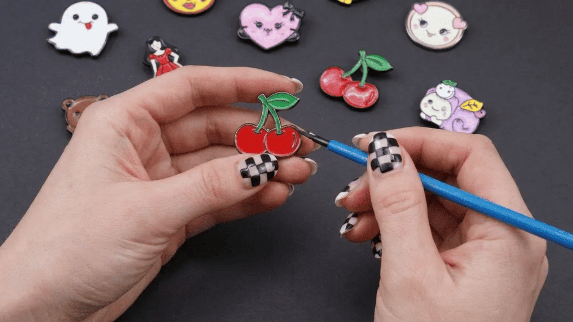 A person applying shiny clear glaze with a paintbrush to a cherry-shaped pin, surrounded by handmade enamel pins on a dark surface.