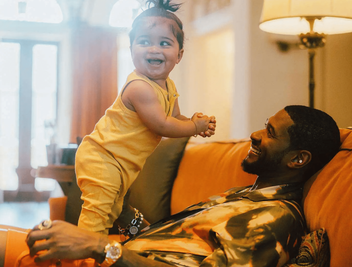 Usher smiling at his youngest son, Sire, standing on his lap in a warmly lit living room.
