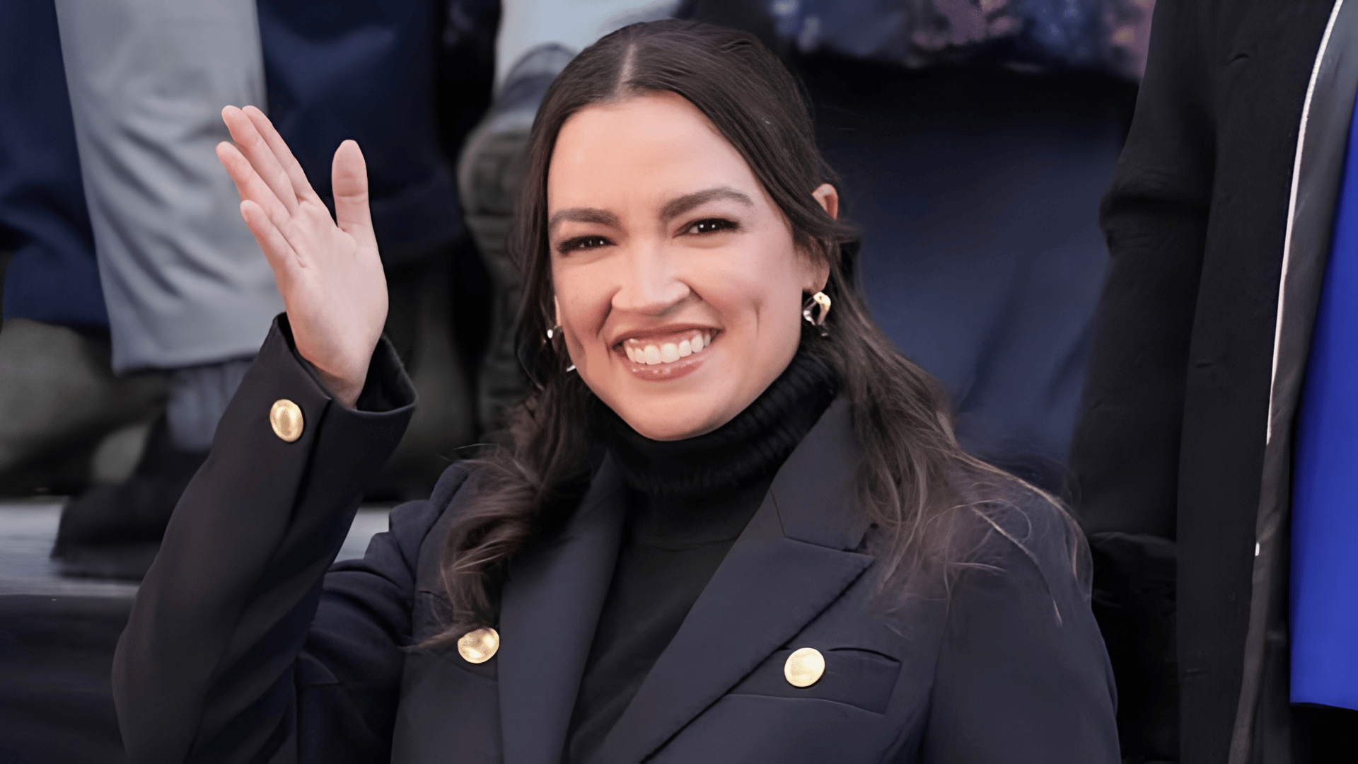 Alexandria Ocasio-Cortez smiling and waving in a dark blazer with gold buttons.