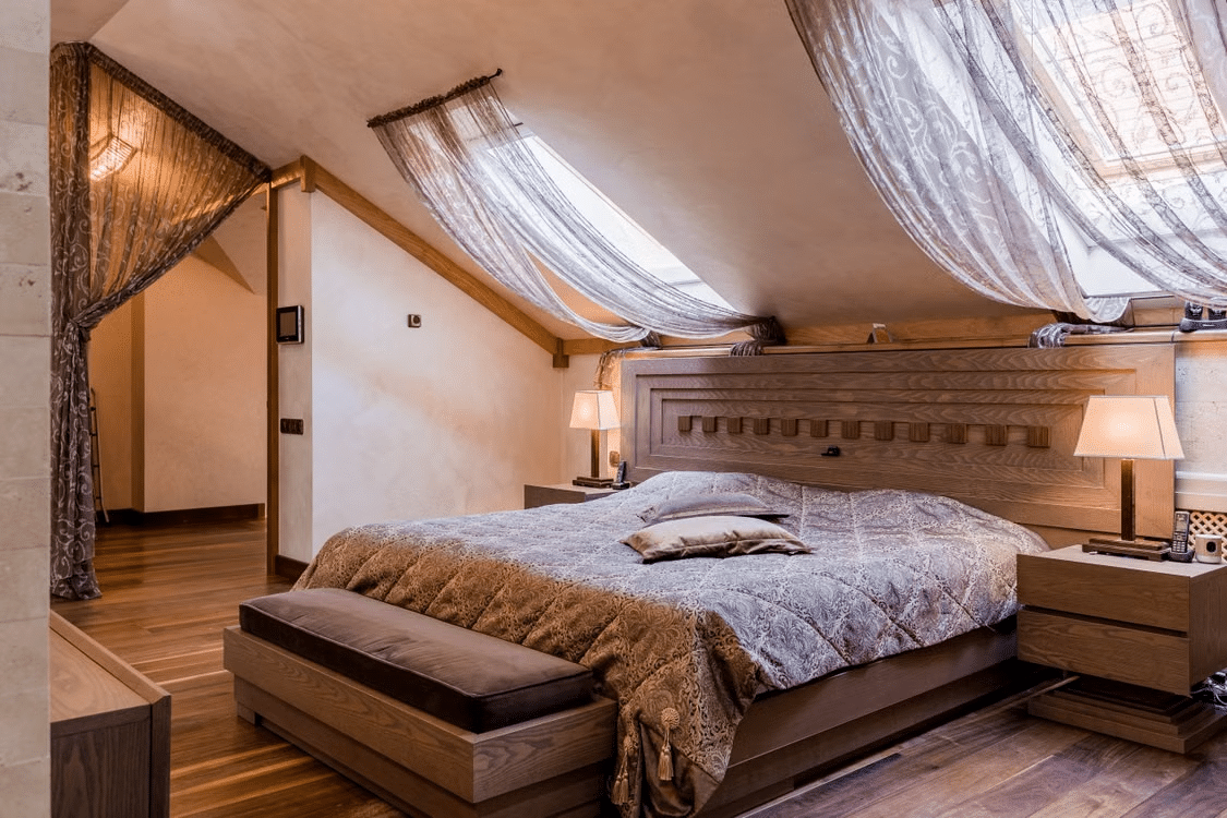Cozy attic bedroom with wooden bed, plush bedding, and sheer draped curtains under warm lighting
