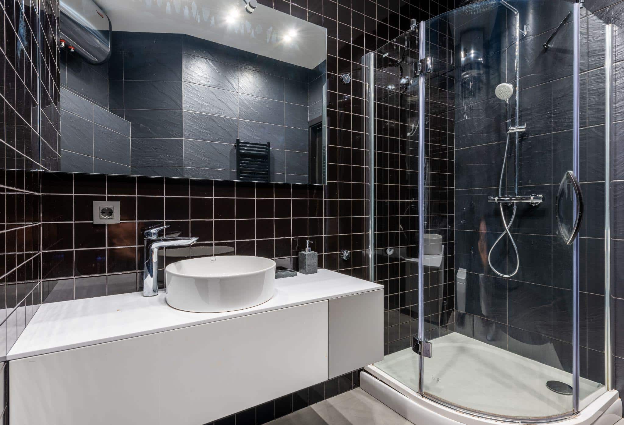Modern bathroom with glass shower enclosure and white countertop against dark tiled walls