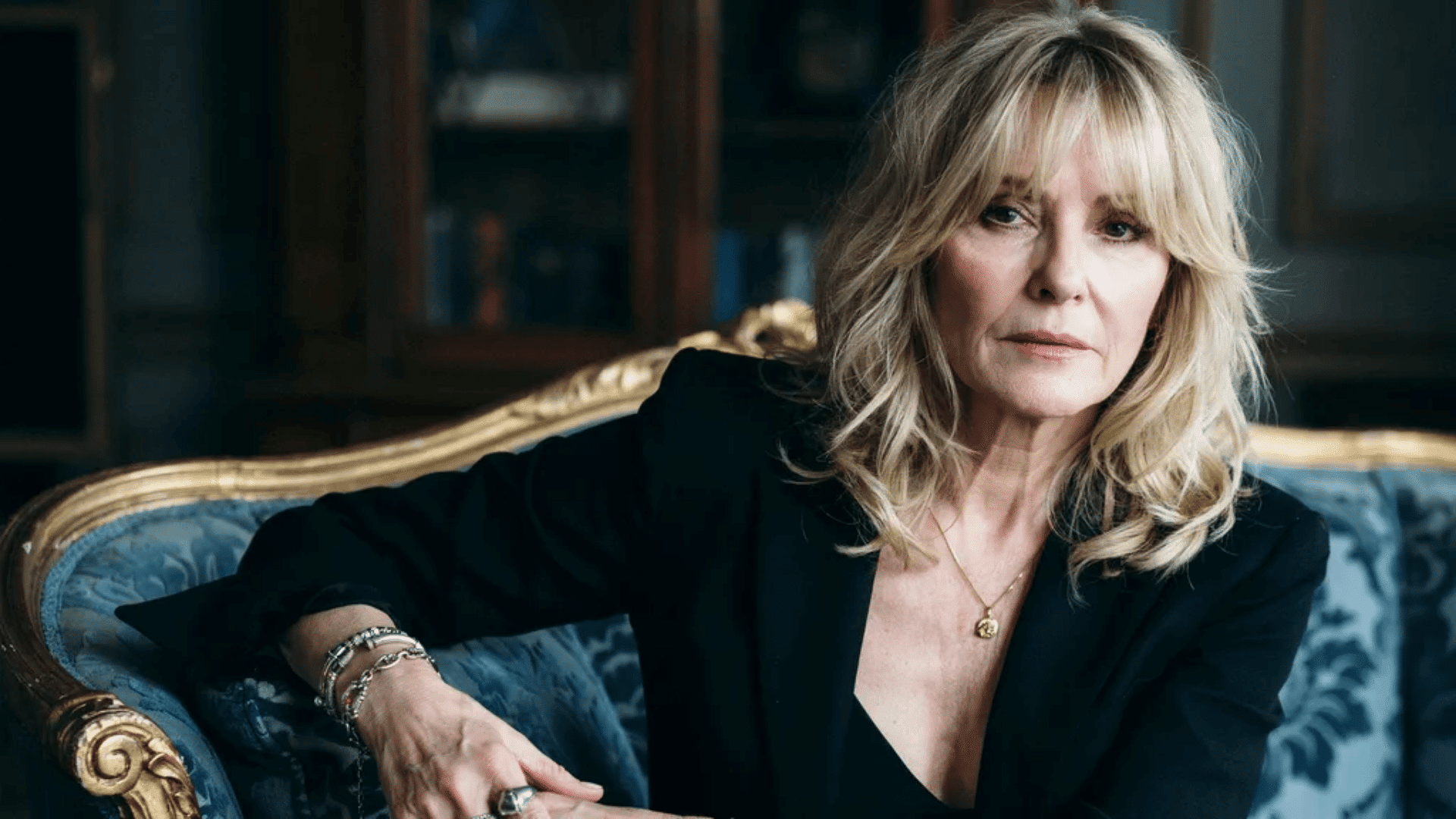 A portrait of Christine McVie wearing a dark blazer and gold necklace, resting her hand on an ornate blue armchair.