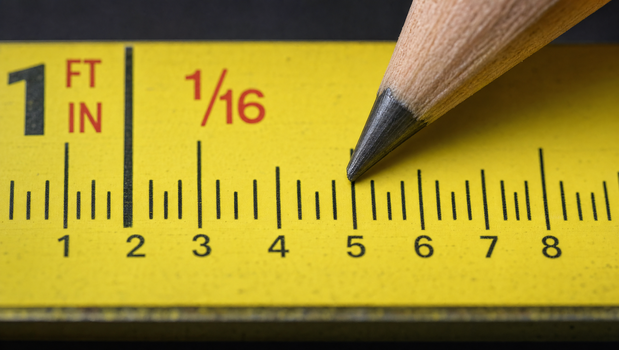 Pencil pointing to small fractional marks on a yellow tape measure, showing how to count 1/16-inch increments.