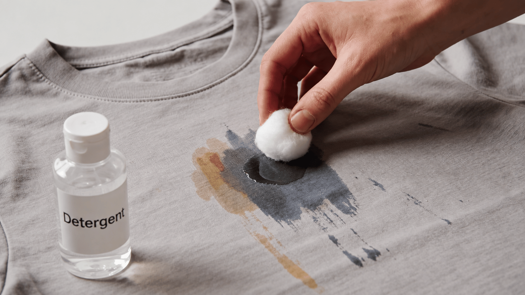 Detergent bottle and cotton pad used to clean a stain on a gray shirt.