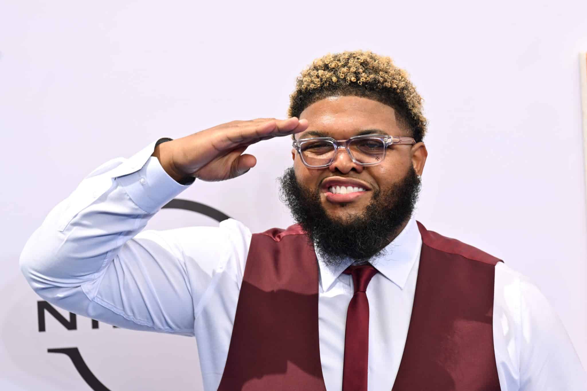 Druski wearing glasses and a burgundy vest while smiling and saluting at an event.