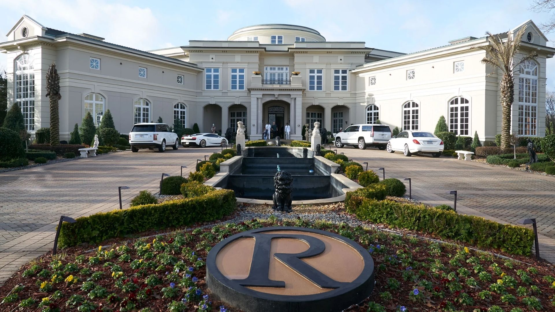 Rick Ross’s mansion with luxury cars, landscaped gardens, and a large “R” emblem