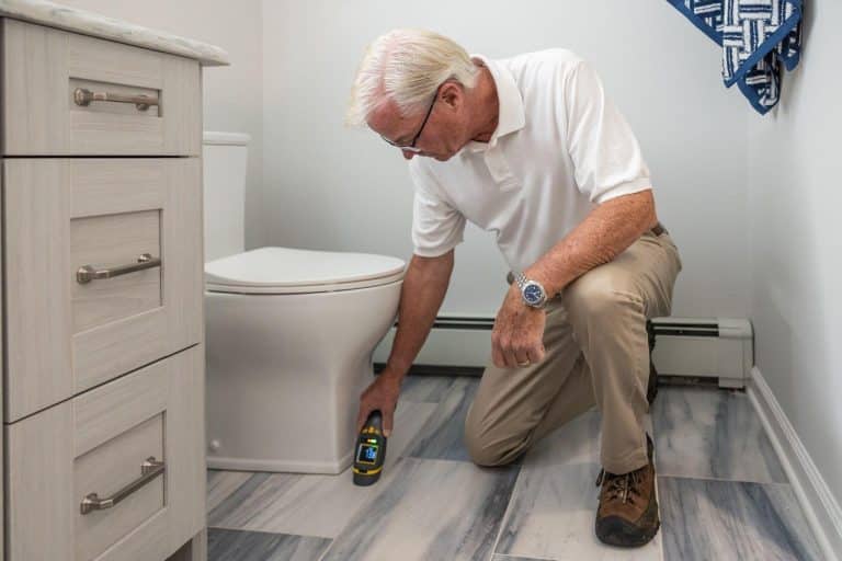 Man kneeling on bathroom floor using moisture meter near toilet in bright, modern bathroom