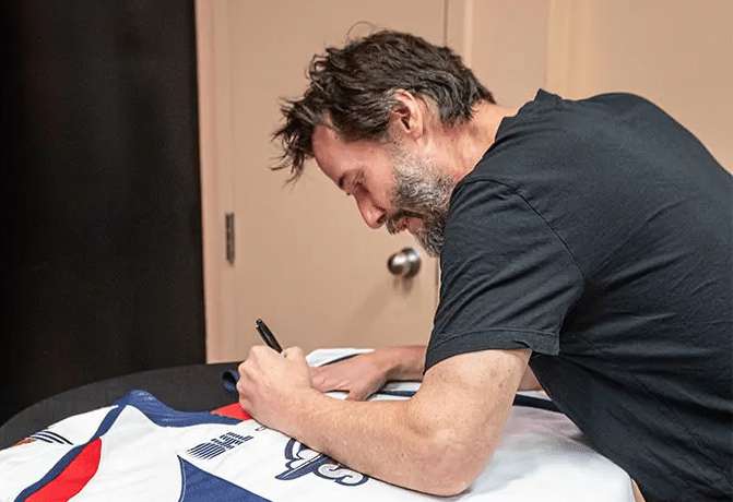 Keanu Reeves leaning over a table to sign an OHL hockey jersey for charity.