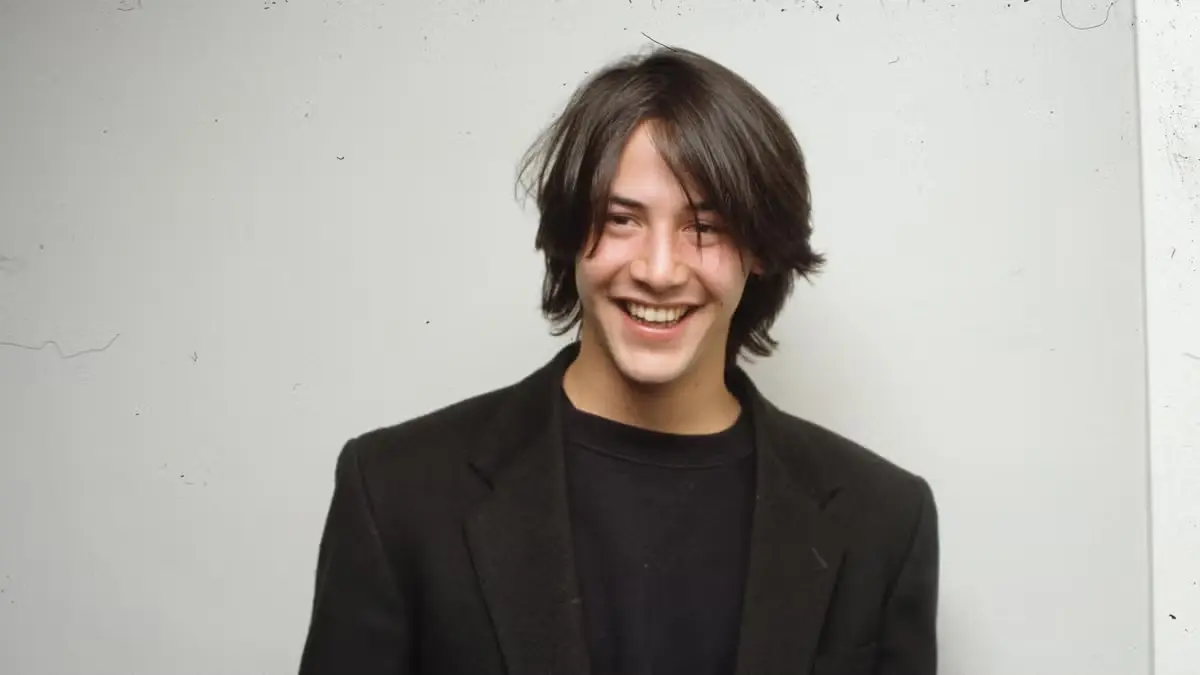Vintage headshot of a young Keanu Reeves smiling with medium-length dark hair in a dark suit jacket.