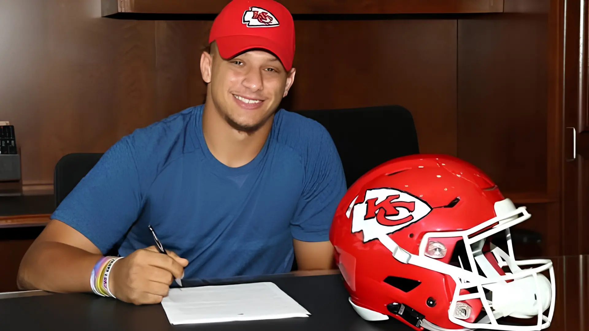 Mahomes in a Kansas City Chiefs cap signing paperwork beside a red Chiefs football helmet.