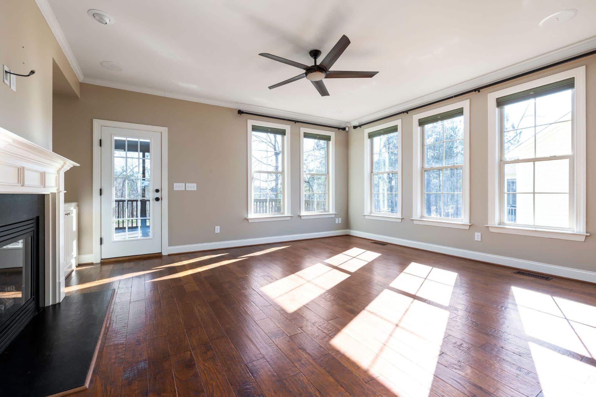 Empty living room with hardwood floors, large windows, and ceiling fan in natural light