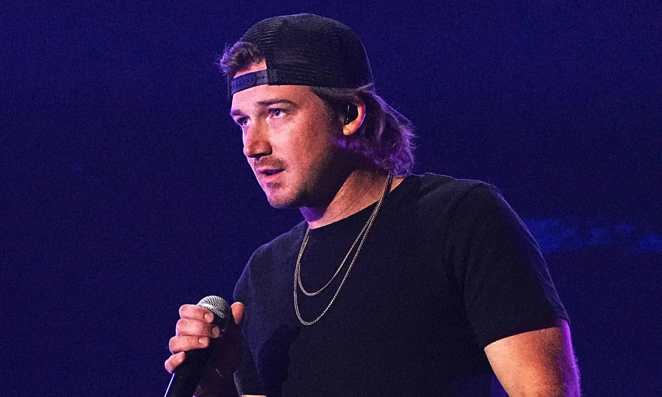 Morgan Wallen performing onstage in a black T-shirt and backward cap while holding a microphone.