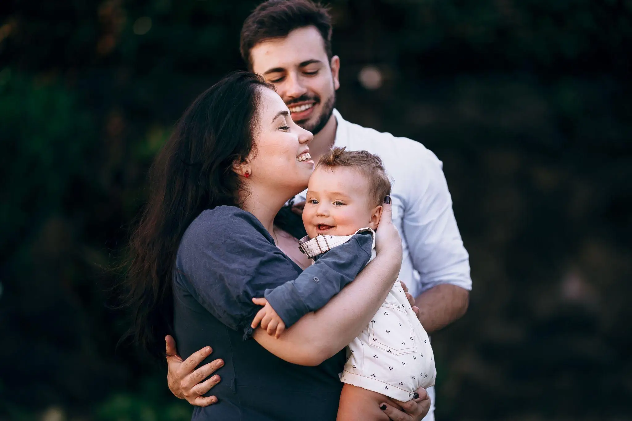 Family embracing with smiling baby in outdoor setting, wearing casual clothing