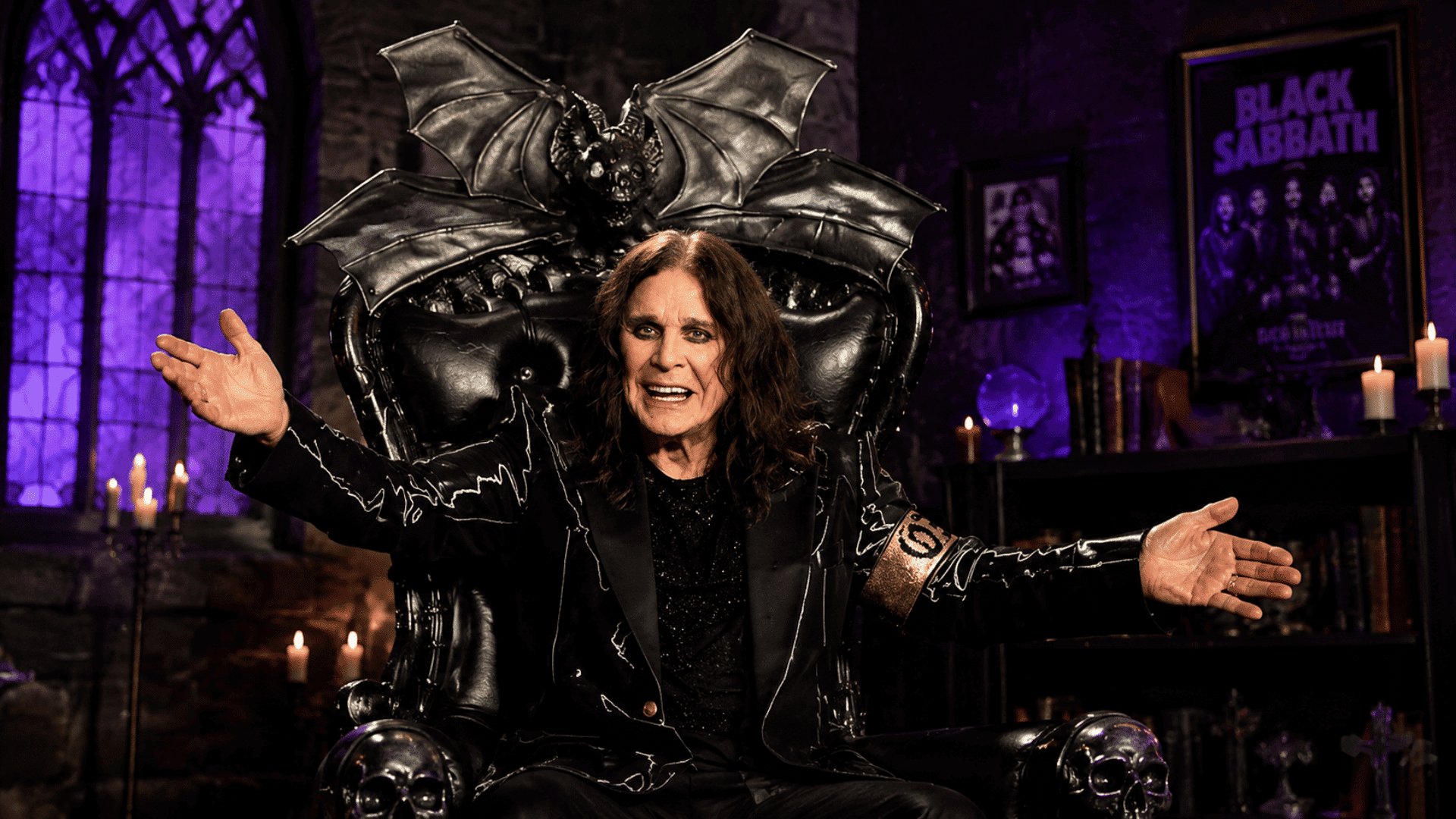 Ozzy Osbourne wearing a long black coat and rings, sitting on a dark ornate throne with his arms outstretched in a gothic-themed room.