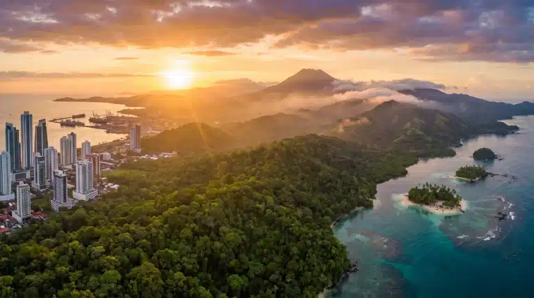 Sunset over coastal city with skyscrapers, lush green forest, and distant mountains by the sea