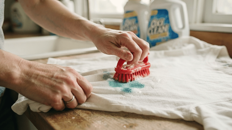 Hands scrubbing a teal stain with a red brush and detergent, showing how to get paint out of clothes fabric.
