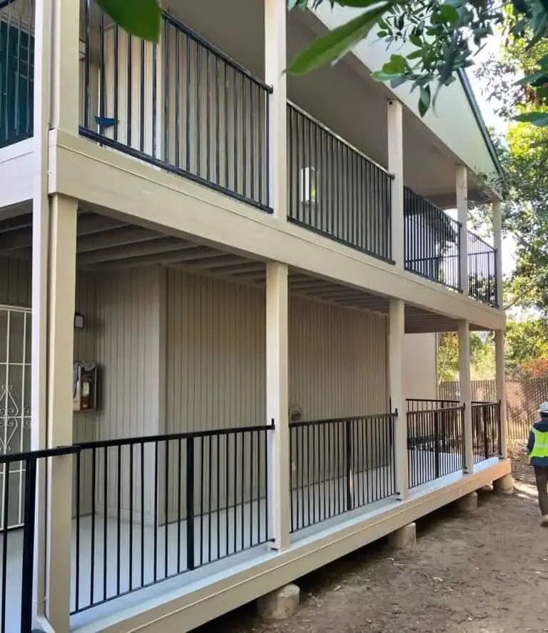 Two-story modular building with beige siding and black railings in a wooded outdoor setting