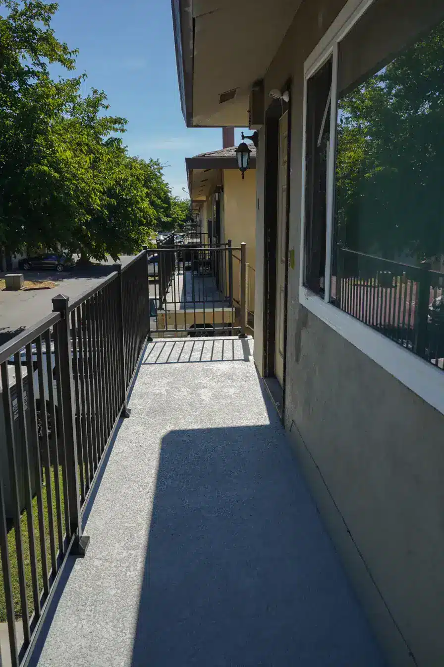 Narrow outdoor walkway with metal railing and tree-lined street under clear blue sky
