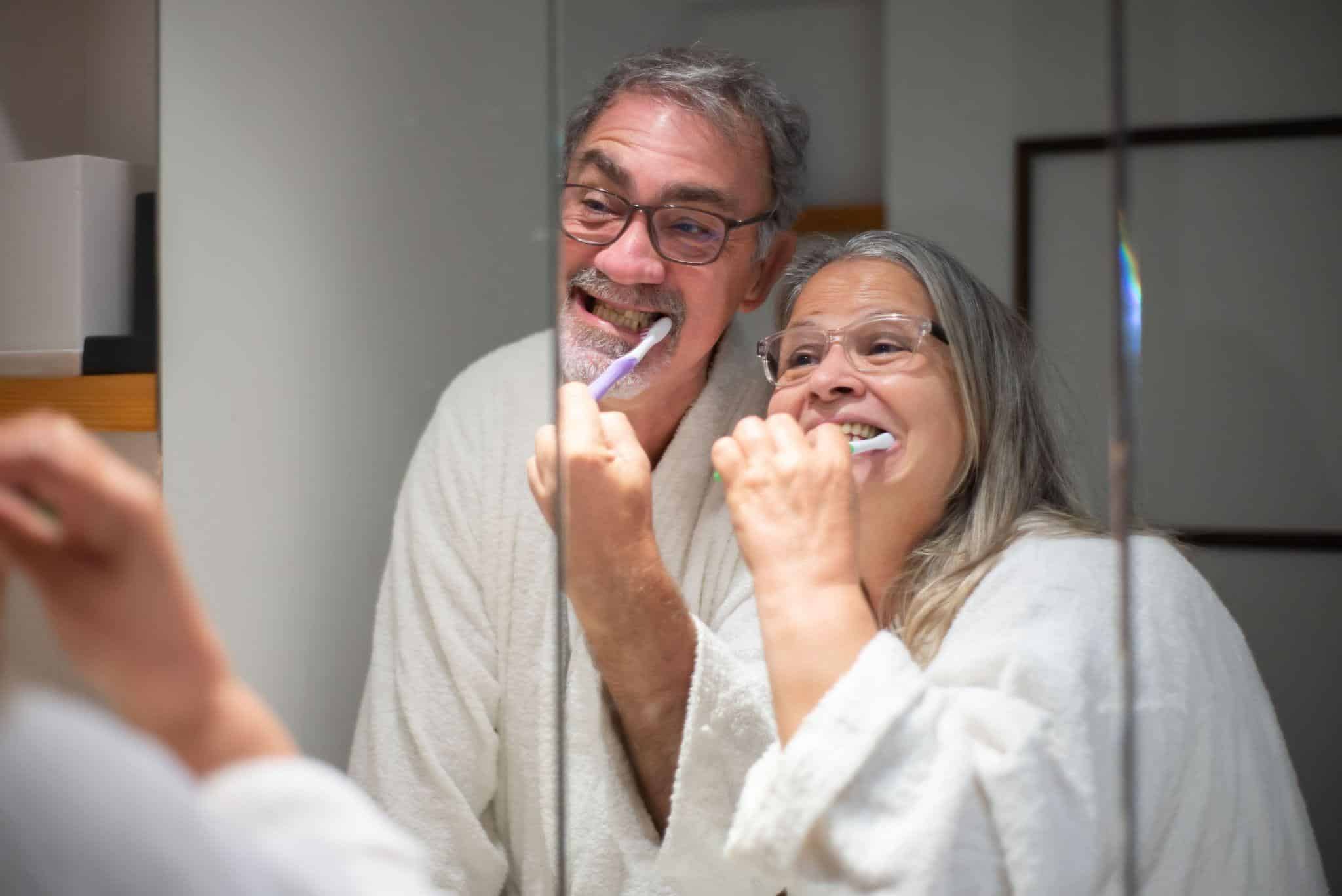 Couple in white robes brushing teeth together in bathroom mirror reflection