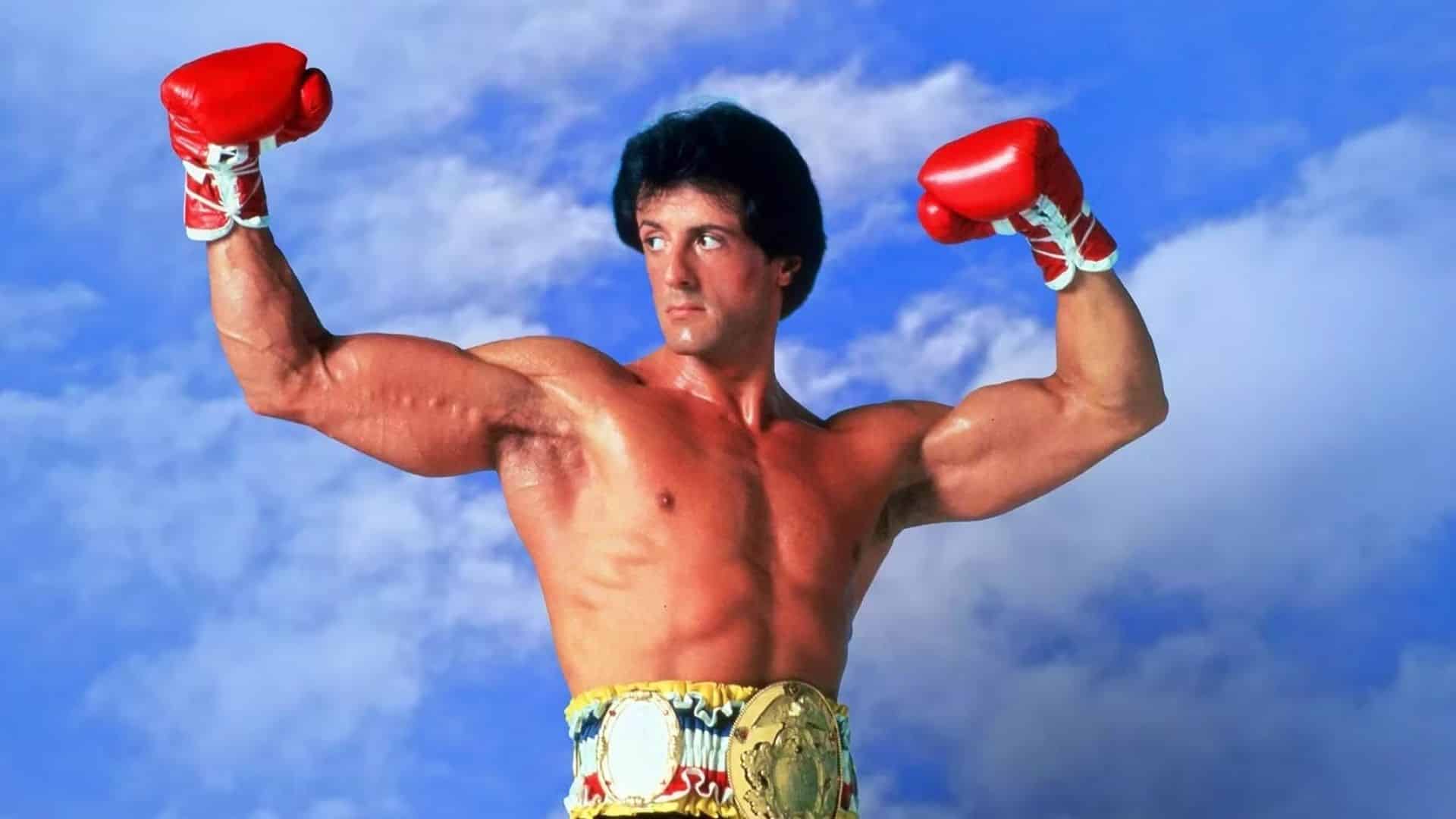 Sylvester Stallone as Rocky Balboa wearing boxing gloves and championship belt in a film scene.