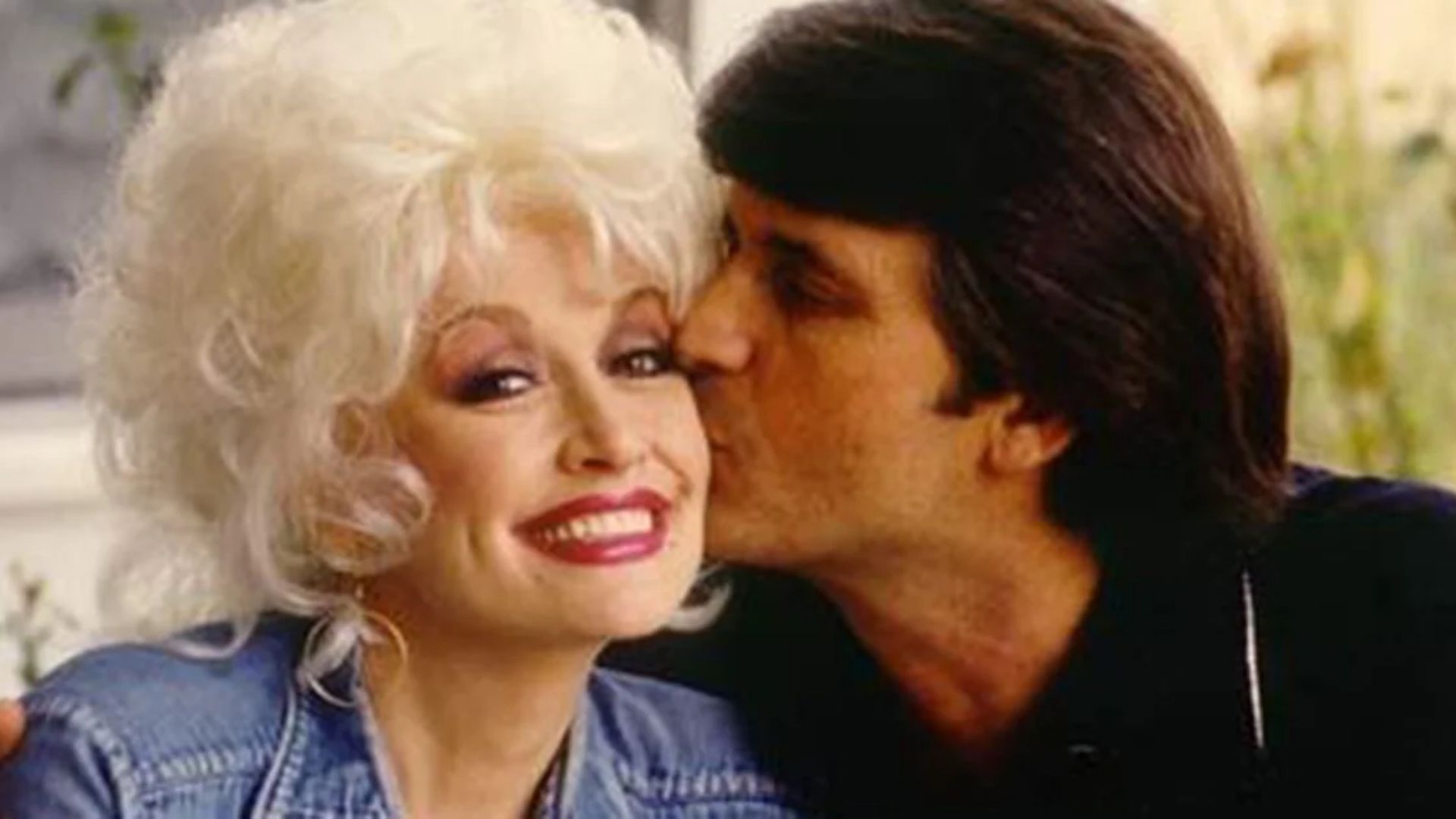 Dolly Parton smiling as her husband, Carl Dean, kisses her cheek.