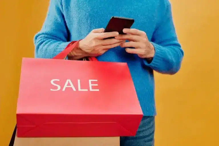 Person in blue sweater holding phone and large red sale shopping bag against yellow background