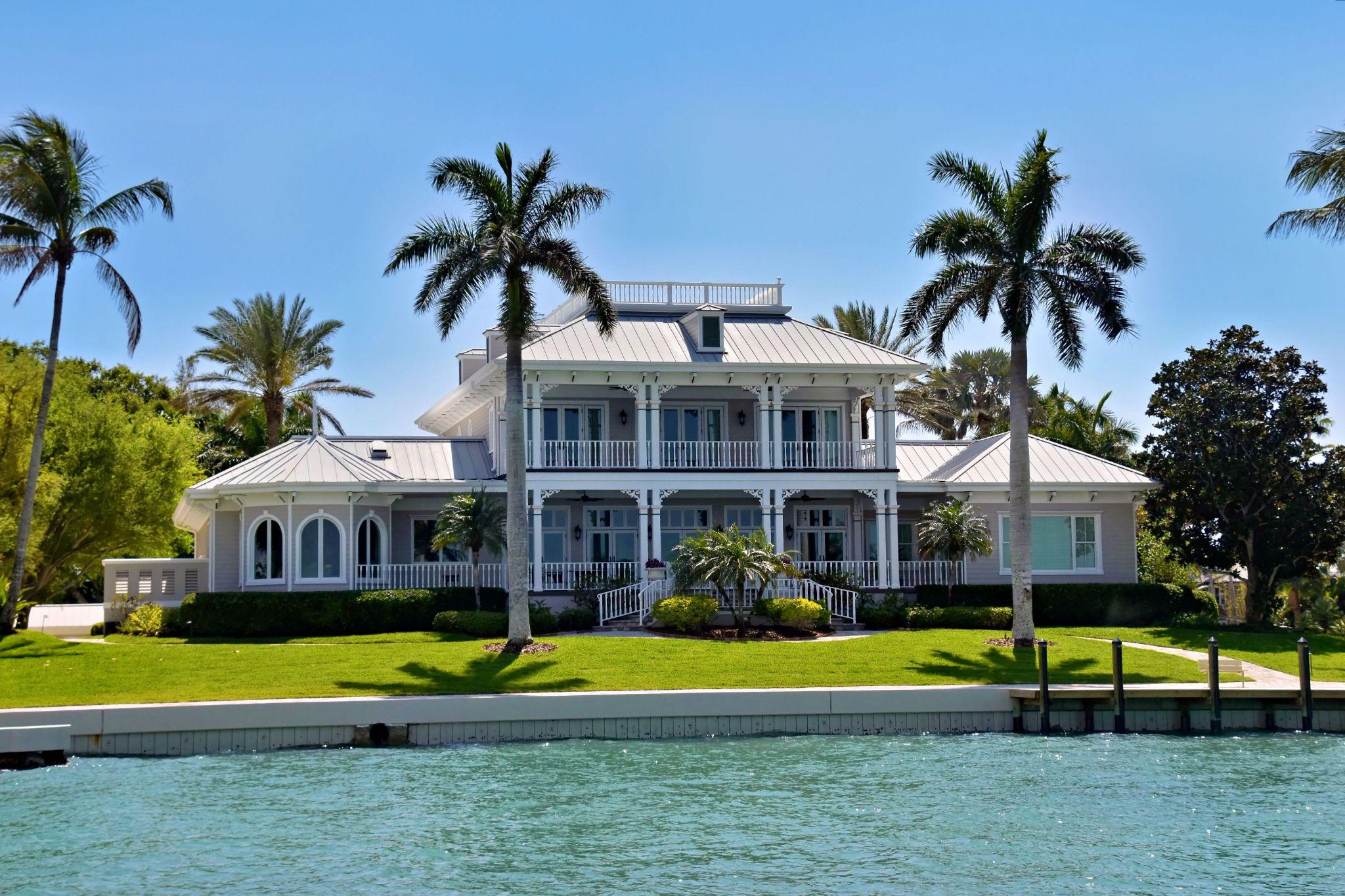 Coastal mansion surrounded by palm trees with water in the foreground under blue sky